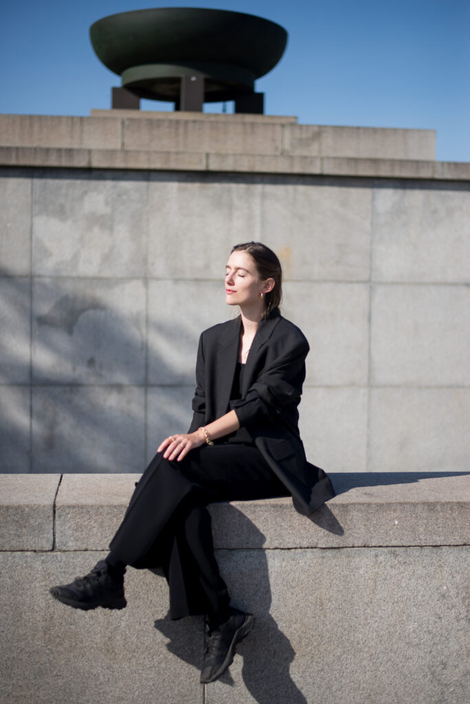 Female model sitting on concrete wall, wearing black blazer.