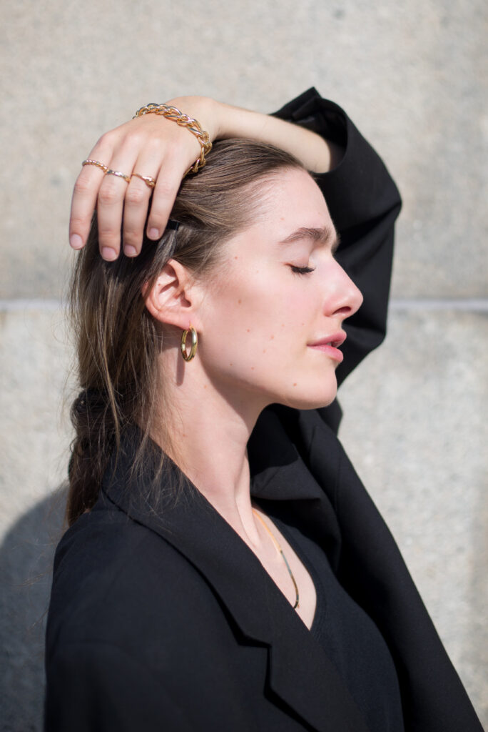 Portrait of blonde female model in front of a concrete wall, wearing black clothing and golden jewellery.