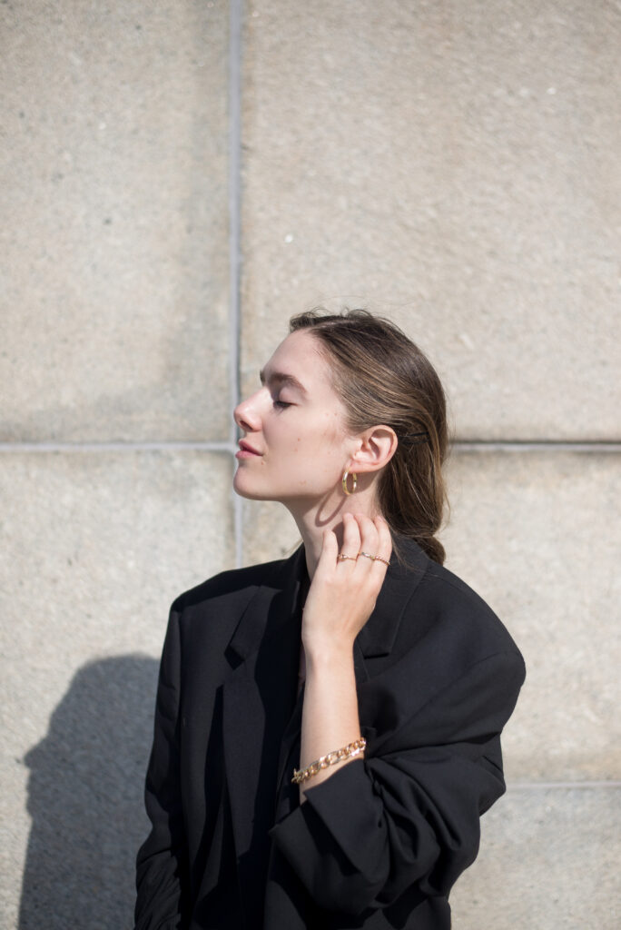 Portrait of blonde female model in front of a concrete wall, wearing black clothing and golden jewellery.