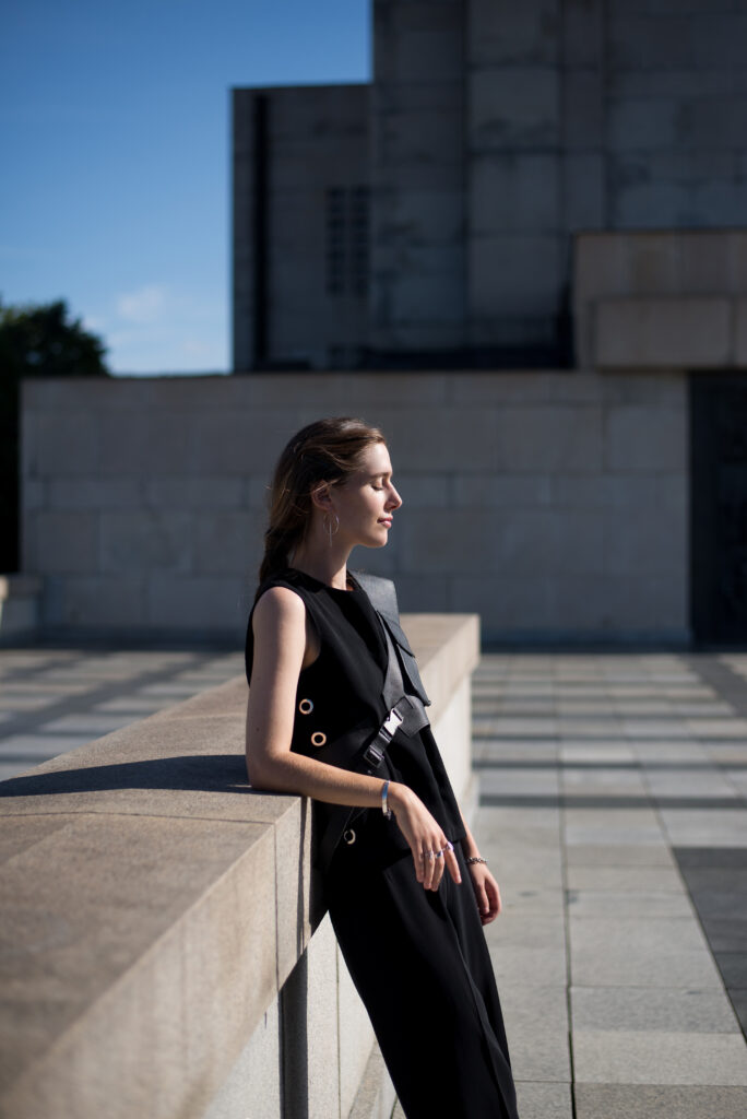 Female model standing in front of monumental building, illuminated by sunlight.