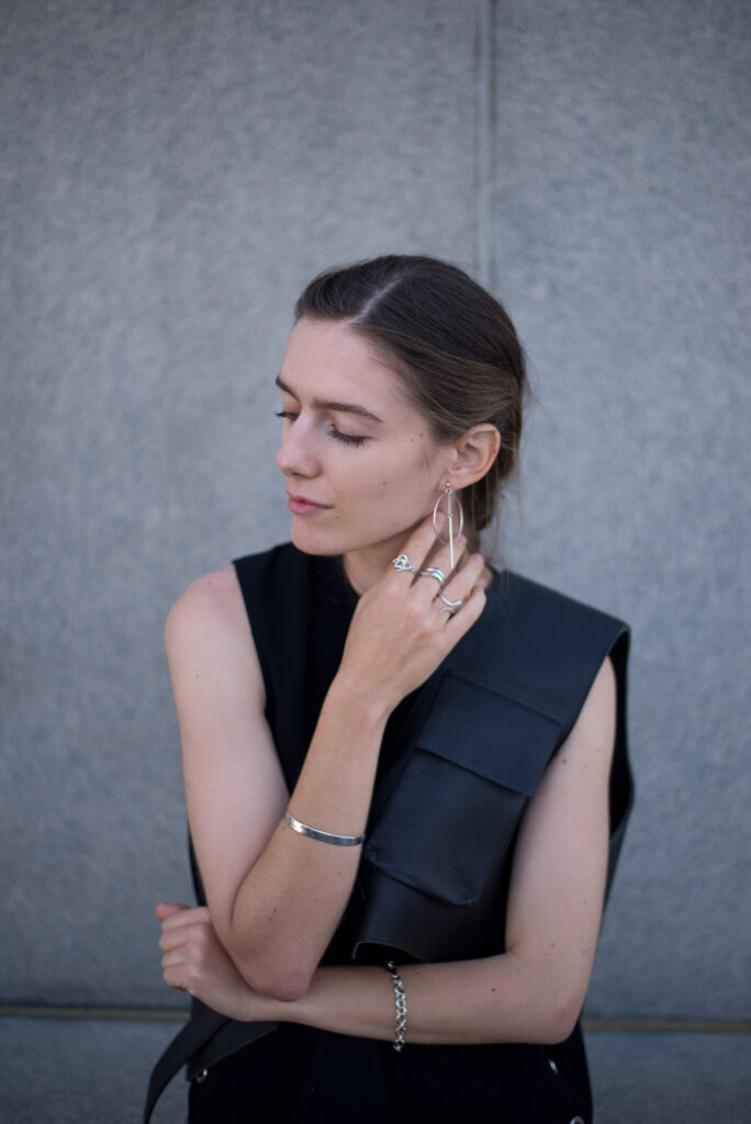 Female model sitting against concrete wall, adorned in silver jewellery.